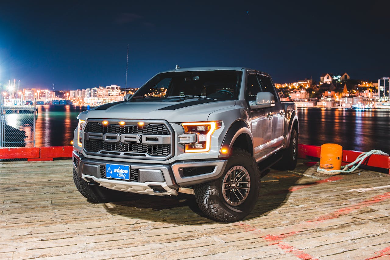 Silver pickup truck parked on a waterfront deck at night with city lights in the background.